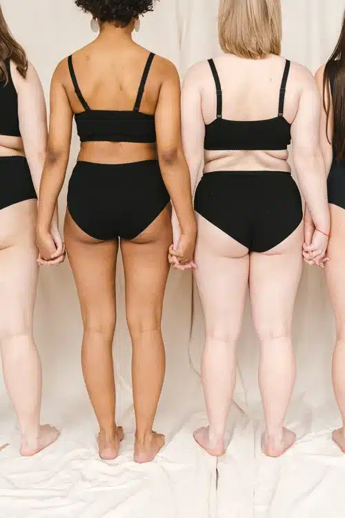 Femmes, corps variés : la diversité corporelle en lingerie. Trois femmes aux corps variés, en sous-vêtements noirs, se tiennent la main, le dos à la caméra. Célébration de la diversité corporelle.