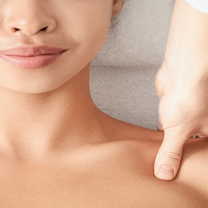 Gros plan d'un soin de massage. Une main applique une pression sur l'épaule d'une femme souriante, mettant en valeur la peau du cou et du décolleté.